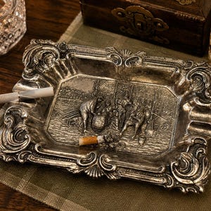 May include: A rectangular silver-toned metal ashtray featuring an ornate, raised border with scrollwork and shell motifs. The center depicts a detailed relief scene of figures gathered around a barrel. A cigarette rests on the edge of the tray.