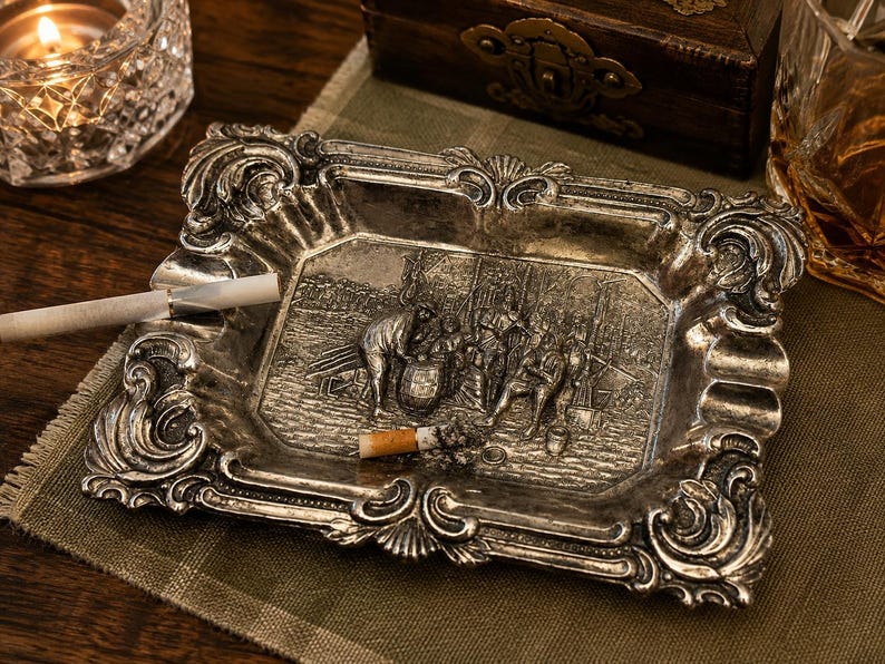 May include: A rectangular silver-toned metal ashtray featuring an ornate, raised border with scrollwork and shell motifs. The center depicts a detailed relief scene of figures gathered around a barrel. A cigarette rests on the edge of the tray.