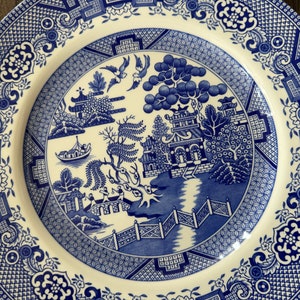 K&ouml;nnte beinhalten: Ein wei&szlig;er Teller aus Keramik mit einem blauen und wei&szlig;en Weidenmuster. Das Muster zeigt eine Br&uuml;cke, einen Fluss, B&auml;ume und eine Pagode.