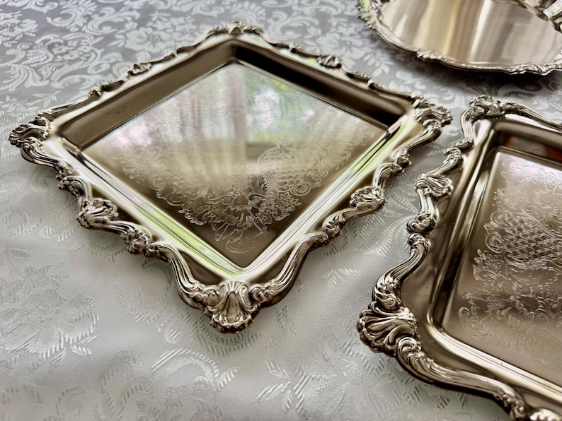 Puede incluir: Un juego de tres bandejas de metal plateado que incluye una bandeja cuadrada, una rectangular y una redonda. Cada bandeja tiene una superficie reflectante con un patr&oacute;n floral grabado y un borde elevado, ornamentado y festoneado.
