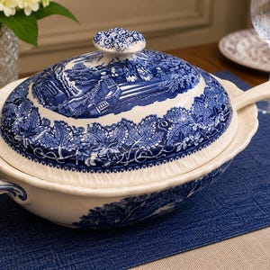 Op de afbeelding: Een witte keramische terrine met een blauw transferware-patroon met een landschapssc&egrave;ne en ingewikkelde bladranden. De serveerschaal met deksel bevat twee handgrepen aan de zijkant en een bijpassende soeplepel met een gebogen steel.