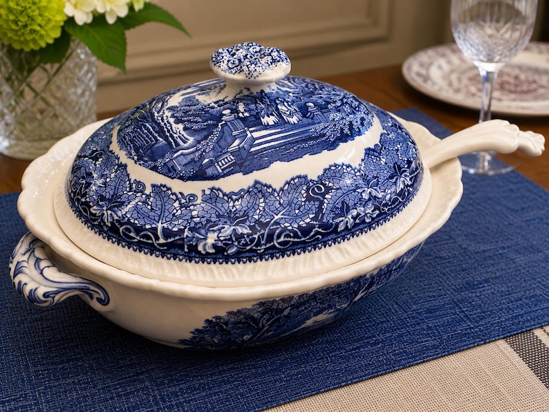 Op de afbeelding: Een witte keramische terrine met een blauw transferware-patroon met een landschapssc&egrave;ne en ingewikkelde bladranden. De serveerschaal met deksel bevat twee handgrepen aan de zijkant en een bijpassende soeplepel met een gebogen steel.