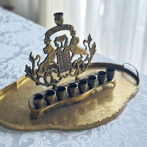 May include: A brass menorah with a decorative design featuring lions and Hebrew text, resting on a matching oval tray. The menorah has eight candle holders and a central shamash holder.