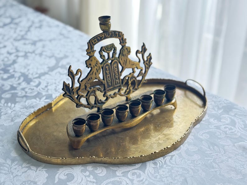 May include: A brass menorah with a decorative design featuring lions and Hebrew text, resting on a matching oval tray. The menorah has eight candle holders and a central shamash holder.