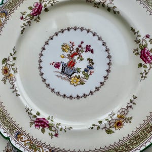 Puede incluir: Un plato de porcelana blanca con un borde verde y dorado. El centro del plato presenta un dise&ntilde;o floral con una peque&ntilde;a planta en maceta en el medio.