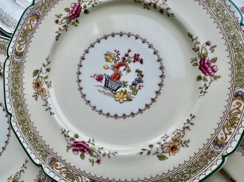 Puede incluir: Un plato de porcelana blanca con un borde verde y dorado. El centro del plato presenta un dise&ntilde;o floral con una peque&ntilde;a planta en maceta en el medio.