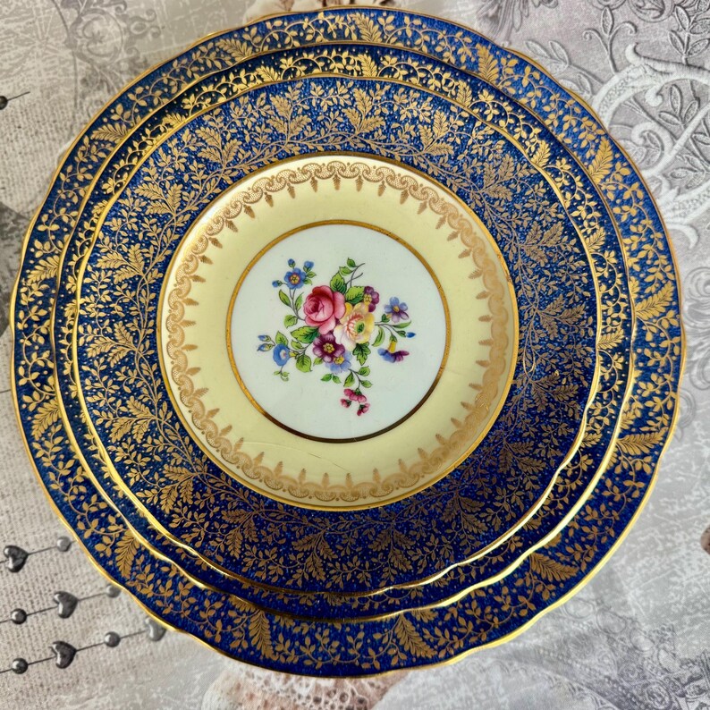 May include: A stack of three antique porcelain plates. The plates feature a navy blue and gold leaf pattern on the outer rim. The center plate has a pale yellow background with a floral bouquet design. The plates are arranged in a nested fashion.