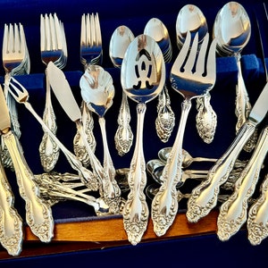 May include: A complete set of ornate silver-colored flatware, including forks, knives, spoons, and serving utensils, arranged in a blue velvet-lined wooden case. The silverware features detailed floral patterns.