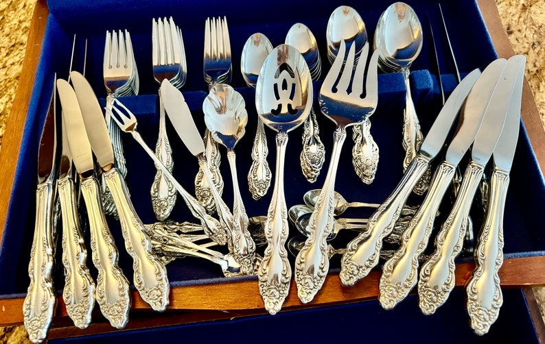 May include: A complete set of ornate silver-colored flatware, including forks, knives, spoons, and serving utensils, arranged in a blue velvet-lined wooden case. The silverware features detailed floral patterns.