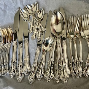 May include: A collection of silver-toned flatware featuring ornate, embossed floral detailing on the handles. The set includes multiple spoons, forks, and knives with polished surfaces, arranged together on a textured, light-colored surface.