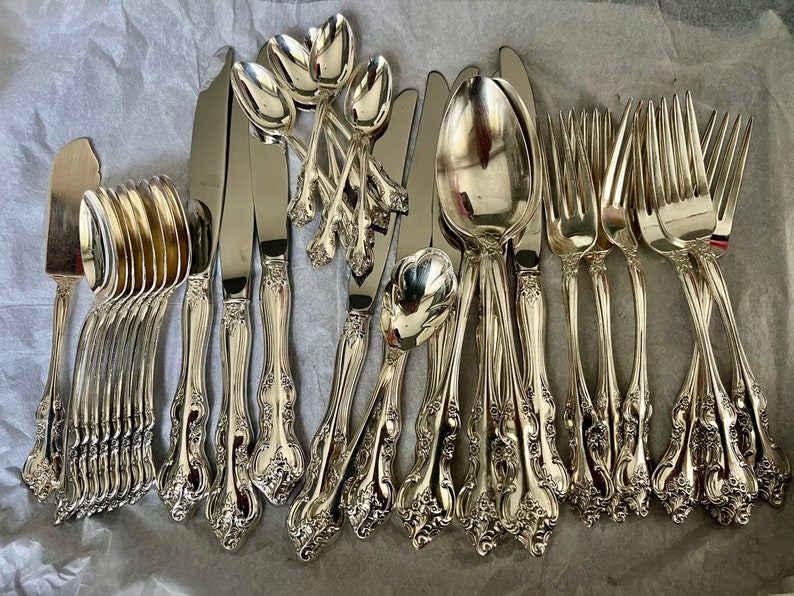 May include: A collection of silver-toned flatware featuring ornate, embossed floral detailing on the handles. The set includes multiple spoons, forks, and knives with polished surfaces, arranged together on a textured, light-colored surface.