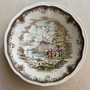 Pode incluir: Uma tigela redonda de cer&acirc;mica com borda recortada e uma moldura de treli&ccedil;a castanha. O centro apresenta uma ilustra&ccedil;&atilde;o paisag&iacute;stica detalhada de um castelo numa colina, um lago com um pequeno veleiro, &aacute;rvores e edif&iacute;cios em tons de verde, vermelho e castanho.