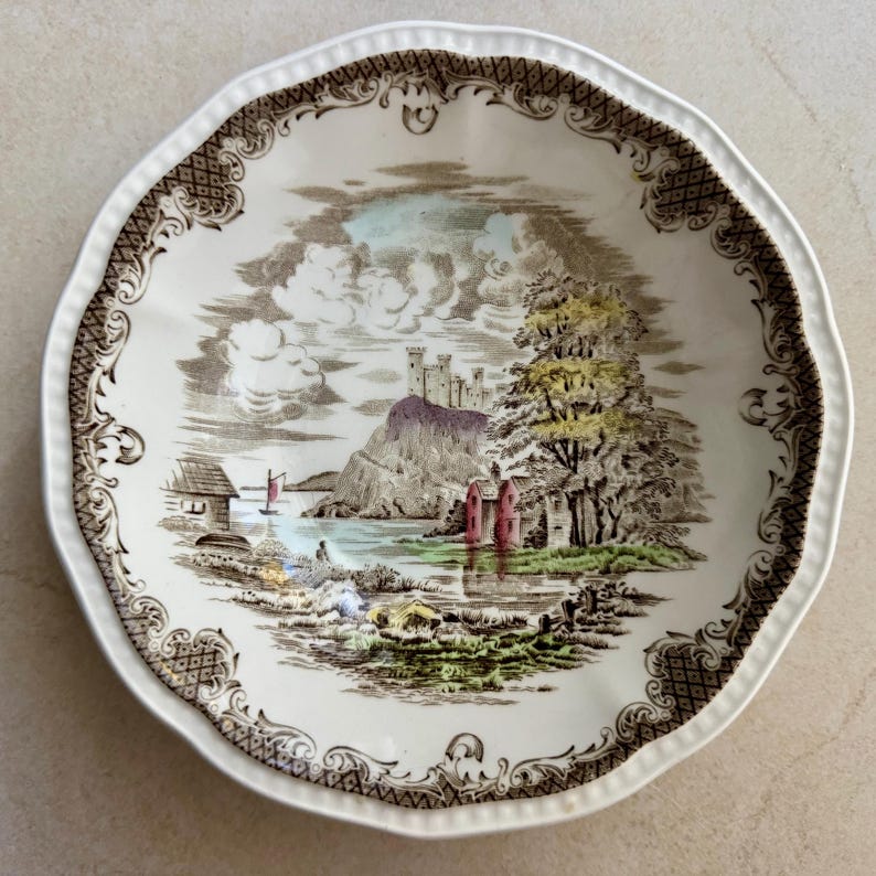 Pode incluir: Uma tigela redonda de cer&acirc;mica com borda recortada e uma moldura de treli&ccedil;a castanha. O centro apresenta uma ilustra&ccedil;&atilde;o paisag&iacute;stica detalhada de um castelo numa colina, um lago com um pequeno veleiro, &aacute;rvores e edif&iacute;cios em tons de verde, vermelho e castanho.
