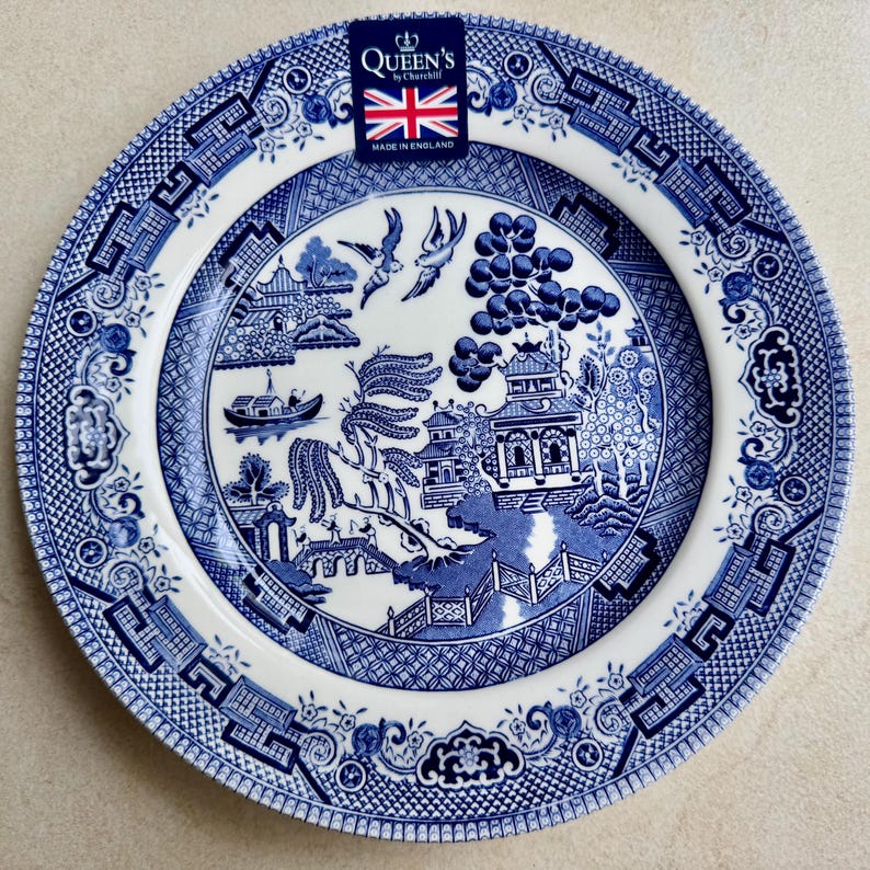 May include: A round ceramic plate featuring a blue and white willow pattern with a central scene of a pagoda, a bridge, and birds. A rectangular label at the top displays the text Queen's by Churchill with a Union Jack and Made in England.