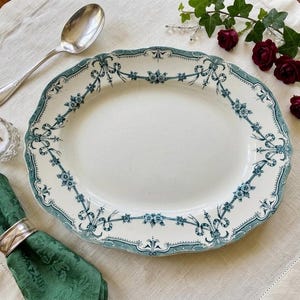 May include: An oval ceramic serving platter featuring a cream-coloured centre and a decorative border with teal blue floral garlands and ribbon motifs. The platter has a scalloped edge and is accompanied by a silver spoon and a green cloth napkin.
