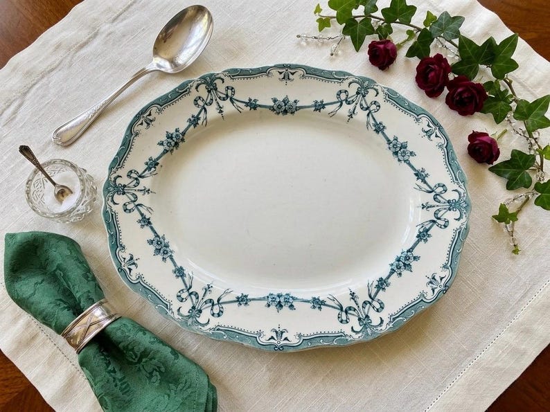 May include: An oval ceramic serving platter featuring a cream-coloured centre and a decorative border with teal blue floral garlands and ribbon motifs. The platter has a scalloped edge and is accompanied by a silver spoon and a green cloth napkin.
