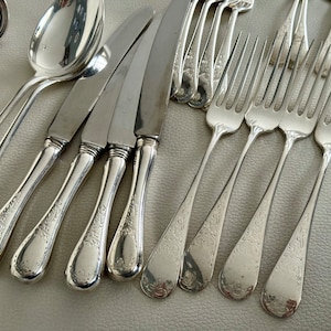 May include: An assortment of antique silverware, including spoons, knives, and forks, arranged on a light-colored surface. The silverware has a shiny, silver finish and features ornate, engraved floral patterns on the handles. The collection includes various sizes of forks and knives.