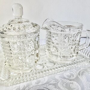 May include: A clear glass sugar bowl with a lid, a matching creamer pitcher with a handle, and a rectangular serving tray. Each piece features a textured hobnail and grid pattern across the surface, crafted from transparent pressed glass.