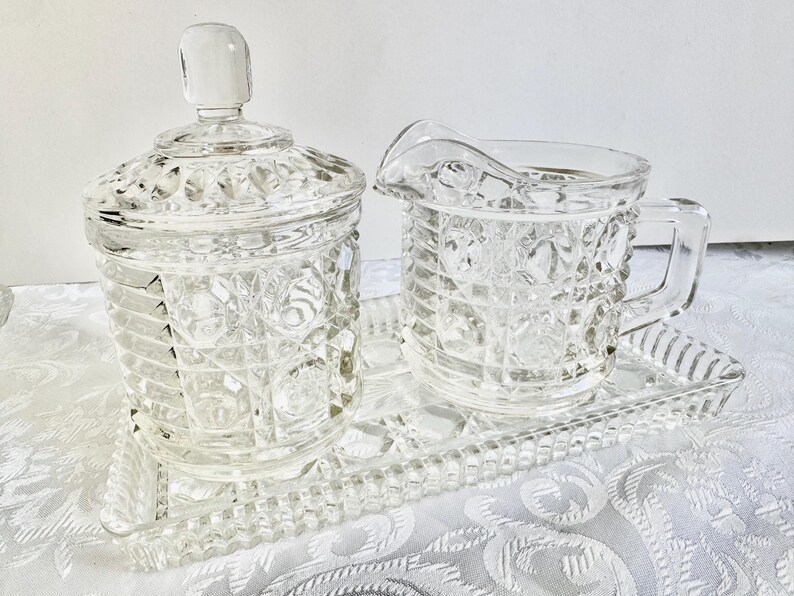 May include: A clear glass sugar bowl with a lid, a matching creamer pitcher with a handle, and a rectangular serving tray. Each piece features a textured hobnail and grid pattern across the surface, crafted from transparent pressed glass.