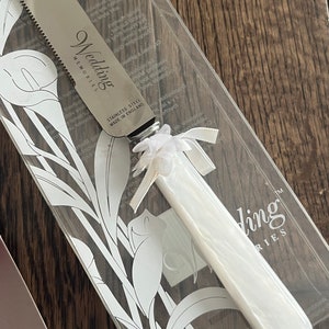 May include: A white wedding cake knife with a silver blade and a white handle. The knife is engraved with the words "Wedding Memories" and "Stainless Steel Made in England".