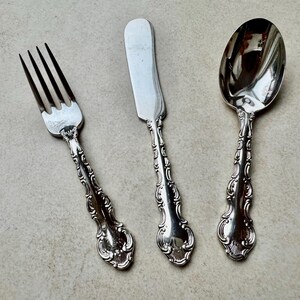 May include: A set of three antique silver-coloured utensils: a fork, a butter knife, and a spoon. Each piece has a decorative, ornate handle with a floral design. The fork has four tines, and the butter knife has a rounded blade.