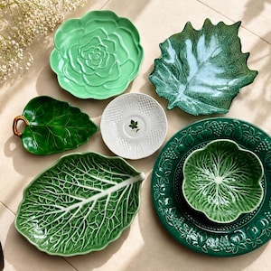 May include: A collection of ceramic tableware including a light green cabbage-shaped plate, a dark green leaf-shaped dish with a brown handle, a white textured saucer, a large dark green leaf plate, and a dark green plate with a matching bowl.