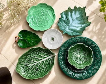 May include: A collection of ceramic tableware including a light green cabbage-shaped plate, a dark green leaf-shaped dish with a brown handle, a white textured saucer, a large dark green leaf plate, and a dark green plate with a matching bowl.