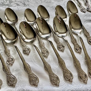 May include: A collection of ornate silver spoons arranged on a white patterned tablecloth. The spoons have a reflective surface and detailed handle designs. The spoons are likely part of a vintage or antique silverware set.