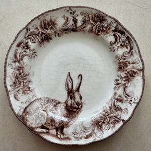 May include: A round ceramic plate with a cream-colored base and a brown transferware pattern. The center features a detailed rabbit illustration, while the scalloped rim is decorated with intricate floral scrolls and roses.