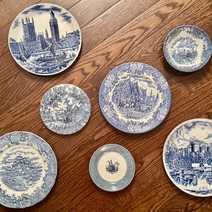 May include: A collection of six round ceramic plates and bowls featuring blue and white transferware patterns. The designs include detailed illustrations of architectural landmarks, rural landscapes, and intricate floral borders on each piece.