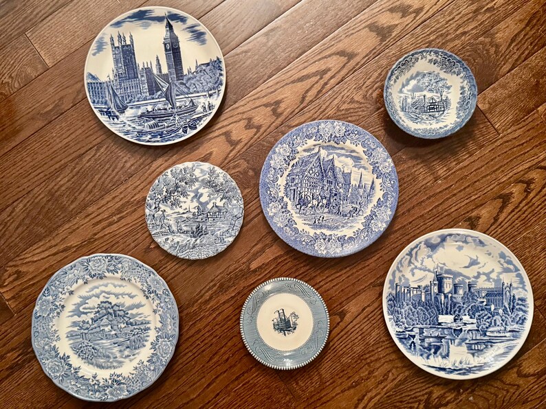 May include: A collection of six round ceramic plates and bowls featuring blue and white transferware patterns. The designs include detailed illustrations of architectural landmarks, rural landscapes, and intricate floral borders on each piece.