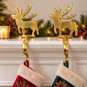 May include: Two gold-toned metal reindeer stocking holders with curved hooks. Each holder features a standing reindeer silhouette with antlers. The hooks extend downward to support a red stocking and a dark green stocking, both with white faux fur cuffs.