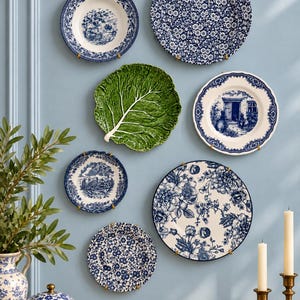 Puede incluir: Una colecci&oacute;n de siete platos decorativos de cer&aacute;mica montados en una pared, con varios patrones florales azules y blancos, escenas de paisajes y un plato con forma de col verde, junto a un jarr&oacute;n azul y blanco y dos candelabros de lat&oacute;n.