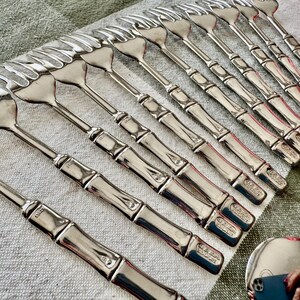 May include: A collection of silver-colored serving forks with a bamboo-style handle design. The forks are arranged on a green and white checkered cloth. The tines of the forks are visible, and the handles have a detailed, textured appearance.