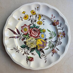 May include: A round, cream-colored ceramic plate with a scalloped edge. The center features a detailed botanical illustration of a bouquet containing pink roses, yellow tulips, small blue flowers, and green leaves with dark stems.