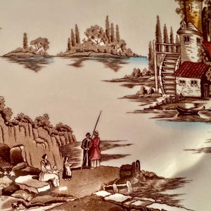 Op de afbeelding: Een keramieken bord met een sepia-kleurige landschapsillustratie van een watermolen, een stenen toren en een meer met eilanden. Op de voorgrond staan figuren bij rotsachtige kliffen en een kleine boot in bruin, wit en blauw.