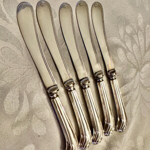 K&ouml;nnte beinhalten: Sechs silberne Buttermesser diagonal angeordnet. Jedes Messer hat eine abgerundete Klinge mit einer zentralen Rille und einem dekorativen Griff. Die Griffe haben ein detailliertes, strukturiertes Design. Die Messer liegen auf einer hellen, gemusterten Oberfl&auml;che.