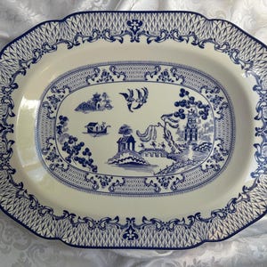 May include: An oval ceramic serving platter featuring a cream-colored base with a blue willow pattern. The design includes a central landscape with a pagoda, bridge, and birds, surrounded by a decorative border of lattice and floral motifs.