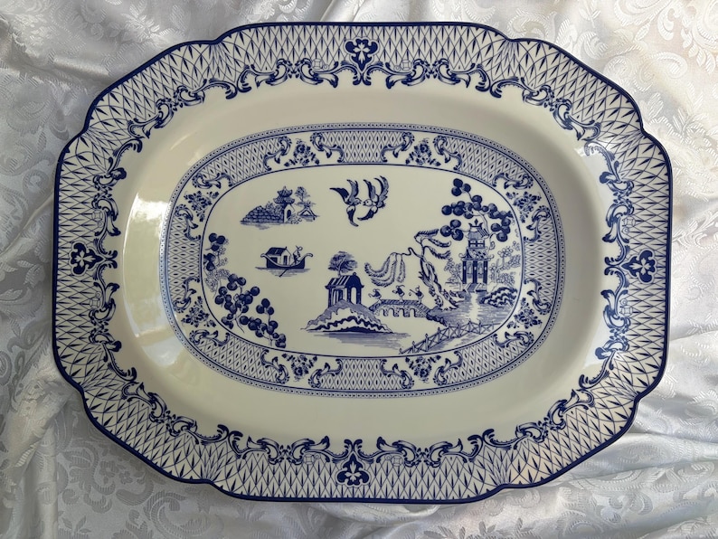 May include: An oval ceramic serving platter featuring a cream-colored base with a blue willow pattern. The design includes a central landscape with a pagoda, bridge, and birds, surrounded by a decorative border of lattice and floral motifs.