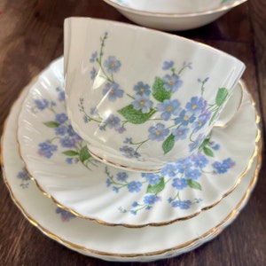 May include: A white teacup, saucer, and plate set with a delicate floral pattern of blue flowers and green leaves. The edges are trimmed with gold. The set is designed for serving tea.