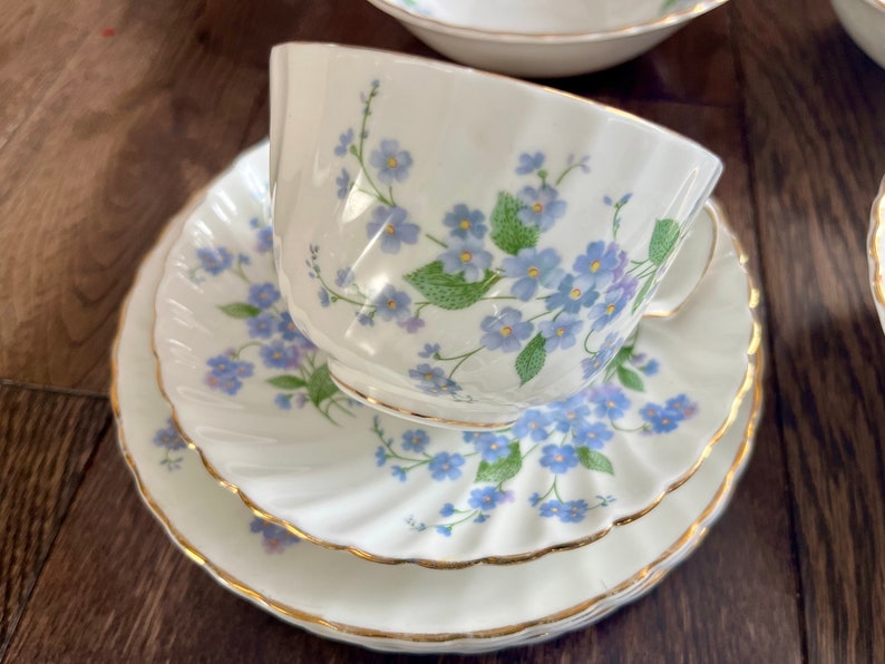 May include: A white teacup, saucer, and plate set with a delicate floral pattern of blue flowers and green leaves. The edges are trimmed with gold. The set is designed for serving tea.