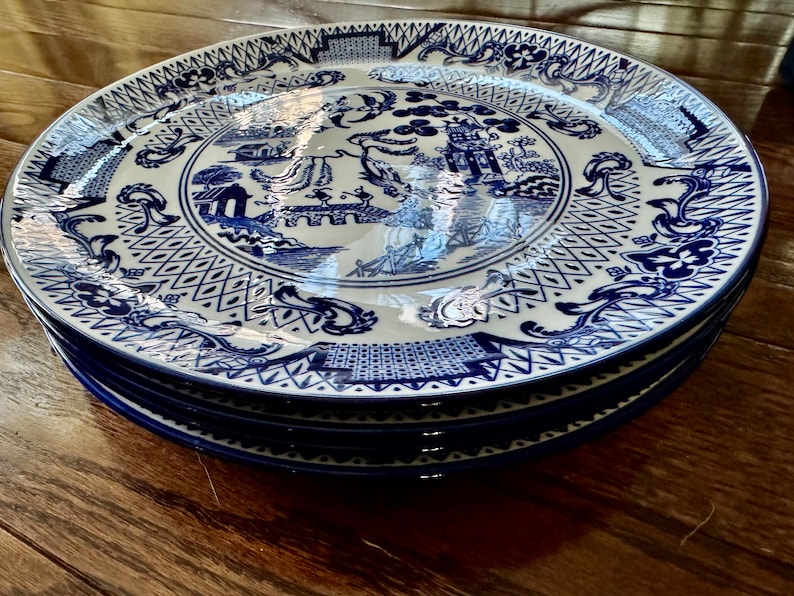 K&ouml;nnte beinhalten: Ein Stapel blau-wei&szlig;er Porzellanteller. Jeder Teller zeigt ein detailliertes, traditionelles, asiatisch inspiriertes Design mit Pagoden, Br&uuml;cken und floralen Mustern. Die Teller sind ordentlich gestapelt und pr&auml;sentieren die komplizierten Kunstwerke und die tiefblaue Farbe.