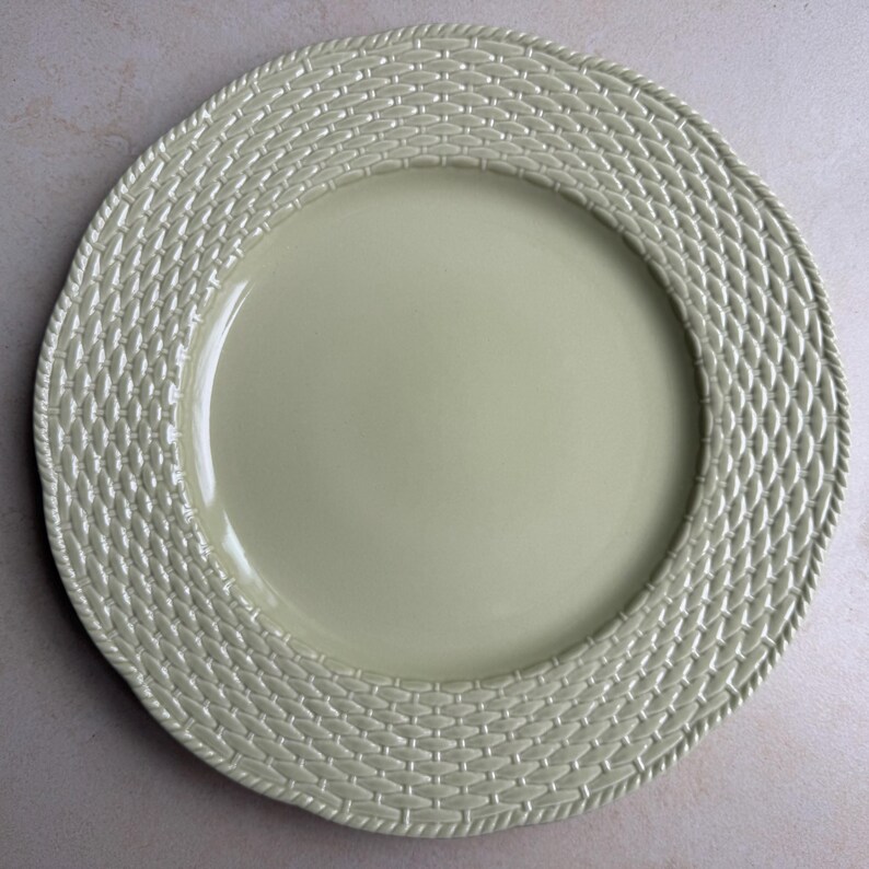 May include: A round, pale sage green ceramic dinner plate featuring a smooth, solid center and a wide, textured rim. The rim displays a raised, repeating basket-weave pattern and is finished with a twisted rope-style border along the outer edge.