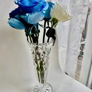 May include: A clear, cut-glass vase with a flared rim and a starburst pattern on the base, containing a bouquet of vibrant blue roses and a single white rose. The glass features intricate geometric facets that catch the light.