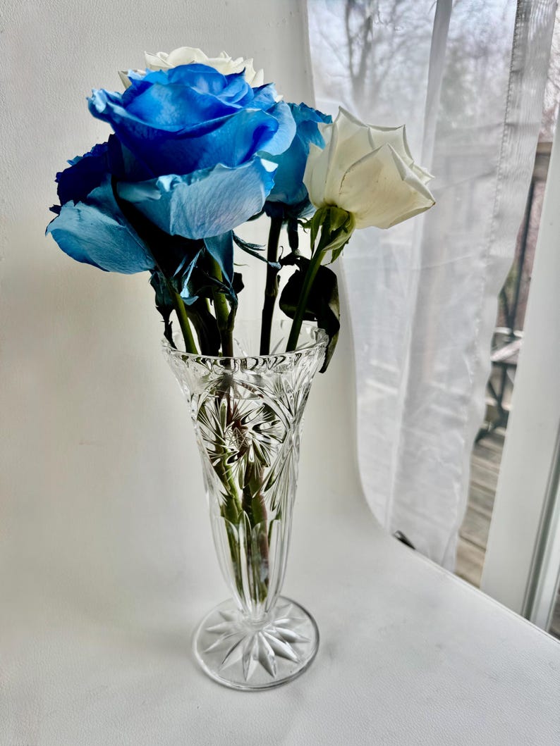 May include: A clear, cut-glass vase with a flared rim and a starburst pattern on the base, containing a bouquet of vibrant blue roses and a single white rose. The glass features intricate geometric facets that catch the light.