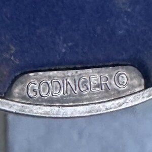May include: A silver-toned metal plaque with a smooth, curved edge featuring the embossed text GODINGER followed by a copyright symbol. The plaque is set against a deep blue, textured surface, highlighting the metallic finish and clear lettering.