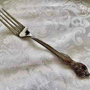 May include: Silverware fork with four tines, featuring an ornate floral design on the handle. The fork is made of a shiny, reflective metal and rests on a white, patterned tablecloth. A classic piece for dining.