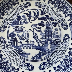 K&ouml;nnte beinhalten: Wei&szlig;e Keramikplatte mit blauem Willow-Muster. Die Platte zeigt eine zentrale Szene mit einer Br&uuml;cke, Figuren und Pagoden, umgeben von einem dekorativen Rand. Die Platte ist rund und hat eine klassische, detaillierte &Auml;sthetik.