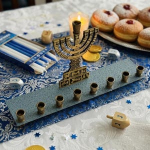 May include: A Hanukkah display with a brass menorah, candles, dreidels, and sufganiyot. The menorah sits on a blue and grey decorative tray. Gold coins and blue and white candles are also present. The table is decorated with blue stars.
