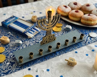 May include: A festive Hanukkah table setting featuring a brass menorah, candles, dreidels, and sufganiyot. The menorah is placed on a blue and gray decorative tray. Gold coins and blue and white candles are also present.